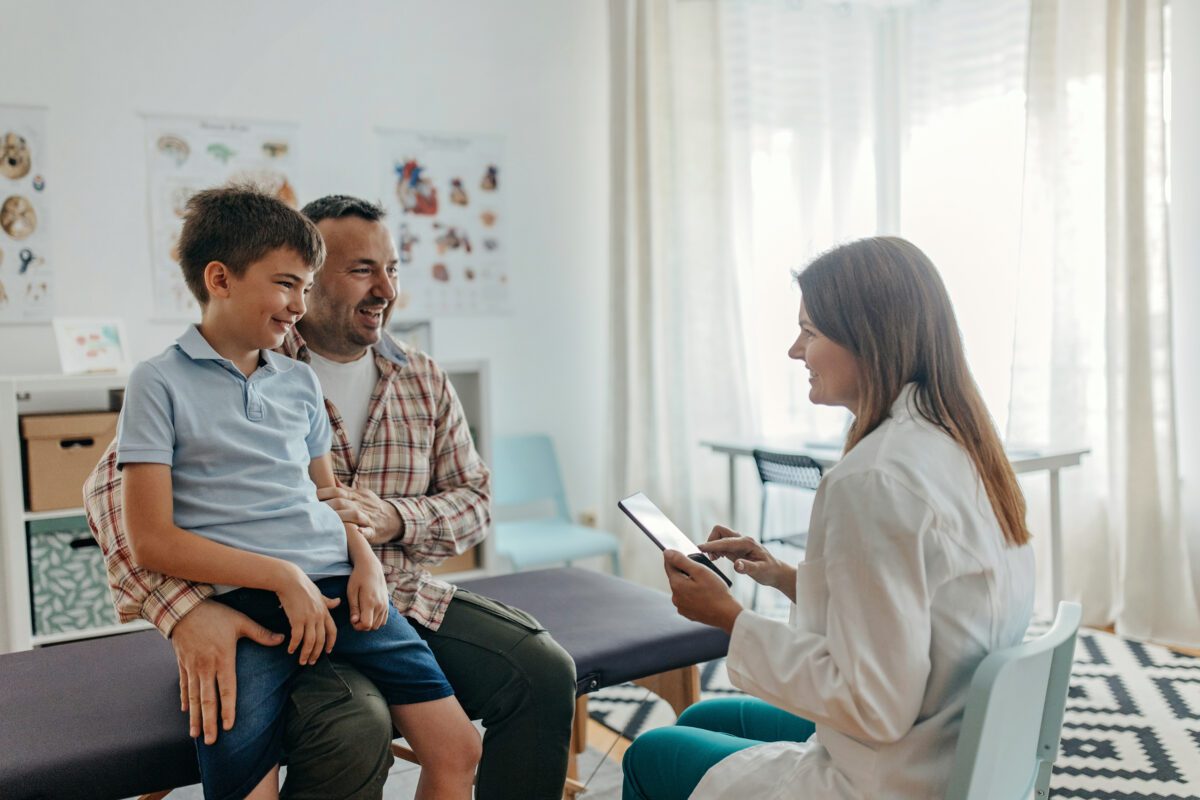 Father and son are unrecognisable, female doctor is examining little boy. She is taking notes on the digital tablet