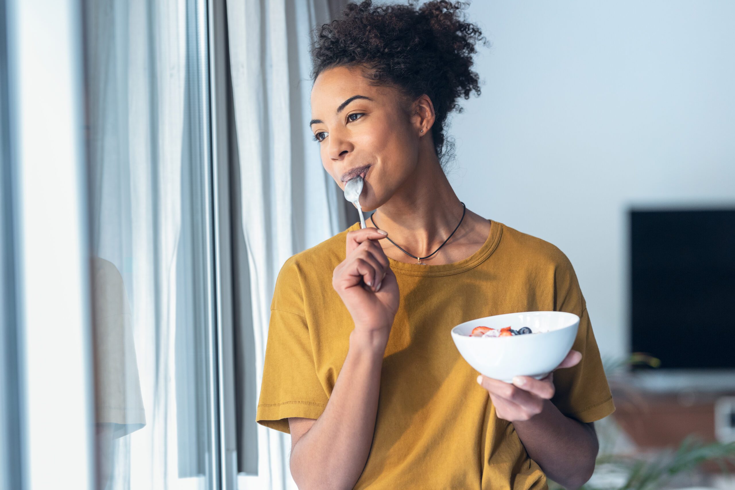 Beautiful mature woman eating cereals and fruits while standing next to the window at home.